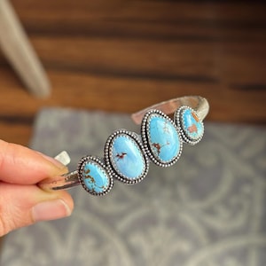 May include: A silver cuff bracelet featuring five oval turquoise stones. Each stone is a vibrant blue with brown matrix patterns, set in a beaded bezel. The bracelet has a hammered texture and is held by a hand.