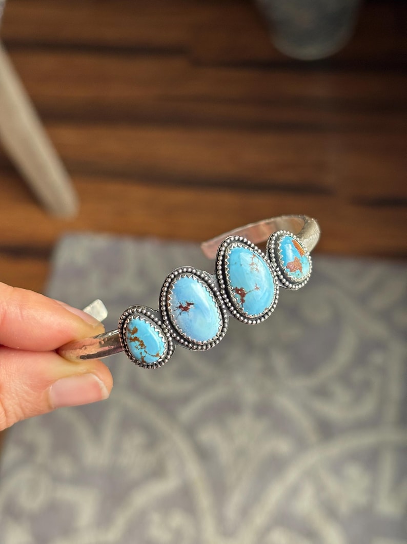 May include: A silver cuff bracelet featuring five oval turquoise stones. Each stone is a vibrant blue with brown matrix patterns, set in a beaded bezel. The bracelet has a hammered texture and is held by a hand.