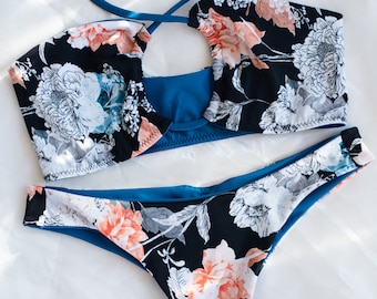 May include: A black and white floral print bikini with a teal blue lining. The top has a halter style with a keyhole cutout in the front. The bottom is a high-cut style.