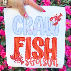 Op de afbeelding: Wit sweatshirt met de woorden "CRAW FISH SEASON" in blauwe en rode letters. Het woord "CRAW" is blauw met een geschulpte rand en verticale strepen. Het woord "FISH" is rood met een geschulpte rand en verticale strepen. Twee rode kreeften zijn ook afgebeeld.