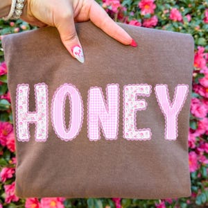 以下が含まれることがあります： 黒い持ち手が付いた白いセラミックマグカップ。黒い太字で「But First, Coffee」と書かれています。ぼやけた背景は、居心地の良い環境を示唆しています。