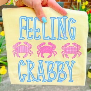 Puede incluir: Una camiseta amarilla clara con las palabras "FEELING CRABBY" en letras azules delineadas. Tres gr&aacute;ficos de cangrejos rosas est&aacute;n debajo de la palabra "FEELING". Los cangrejos tienen diferentes patrones, incluyendo rayas y lunares.