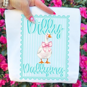 May include: White sweatshirt featuring a cartoon goose with a pink bow and the words "Willy Dallying" in teal. The design is set within a teal and white striped frame, creating a playful and whimsical aesthetic.