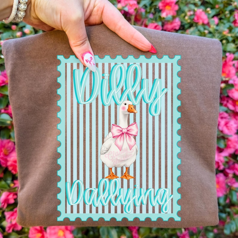 May include: A brown t-shirt featuring a cartoon goose wearing a pink bow, set within a blue and brown stamp-like design. The text "Willy Dallying" is written in a teal script above and below the goose illustration.