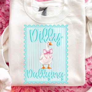 May include: White sweatshirt with a graphic of a goose wearing a pink bow, inside a blue frame with the words "Dilly Dallying". The goose has an orange beak and feet. The background has blue and white vertical stripes.