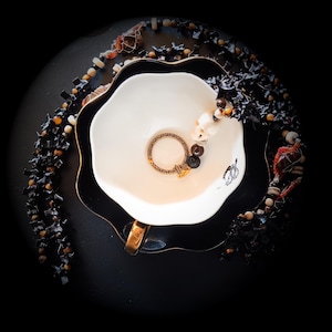 Puede incluir: Una taza y platillo blancos con borde festoneado y ribete dorado, que contiene un anillo dorado y una pulsera de cuentas con un dije de calavera. Un collar de cuentas negro y otras cuentas decorativas rodean la taza sobre una superficie negra.