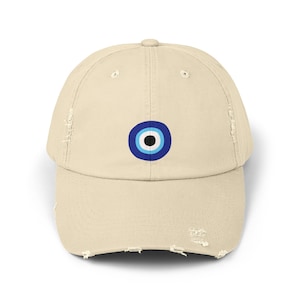 Puede incluir: Una gorra de béisbol desgastada de color beige con un diseño de ojo turco azul y blanco bordado en la parte delantera.