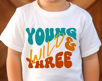 May include: White t-shirt with the text "Young Wild & Three" in colorful, retro-style lettering. The text is in teal, orange, and yellow.