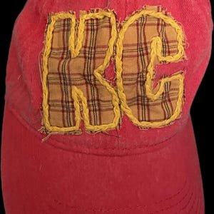 Pu&ograve; includere: Cappellino da baseball rosso con le lettere "RC" applicate sul davanti. Le lettere sono in tessuto a quadri e delineate con cuciture gialle. Il cappellino ha una visiera curva e un bottone sulla parte superiore.