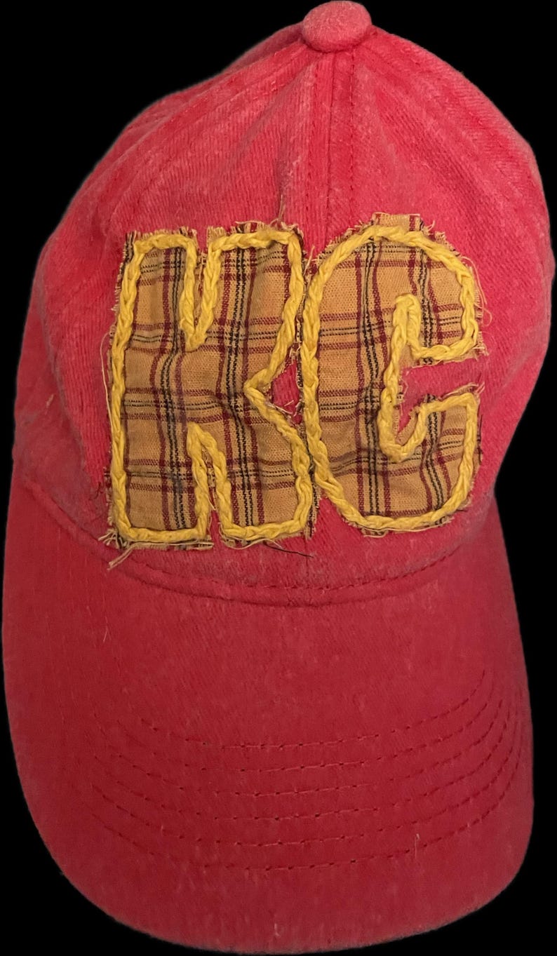 Pu&ograve; includere: Cappellino da baseball rosso con le lettere "RC" applicate sul davanti. Le lettere sono in tessuto a quadri e delineate con cuciture gialle. Il cappellino ha una visiera curva e un bottone sulla parte superiore.