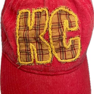 Puede incluir: Gorra de béisbol roja con las letras "KC" aplicadas en la parte delantera. Las letras son de tela de cuadros y están delineadas con costuras amarillas. La gorra tiene una visera curva y un botón en la parte superior.