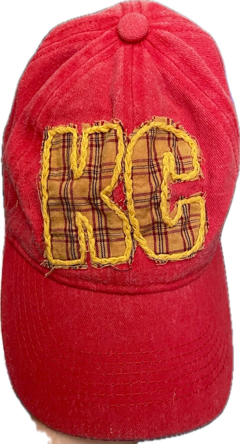 Pu&ograve; includere: Cappellino da baseball rosso con le lettere "KC" applicate sul davanti. Le lettere sono in tessuto a quadri e delineate con cuciture gialle. Il cappellino ha una visiera curva e un bottone sulla parte superiore.