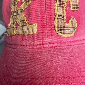 Pu&ograve; includere: Un cappellino da baseball rosa con le lettere "KC" applicate sul davanti. Le lettere sono realizzate in tessuto a quadri con cuciture gialle. Il cappellino &egrave; realizzato in un materiale morbido e strutturato.