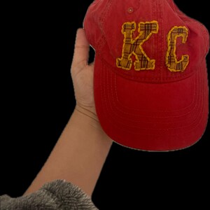 Pu&ograve; includere: Cappellino da baseball rosso con le lettere "KC" in tessuto a quadri giallo. Il cappellino &egrave; tenuto su uno sfondo nero. Ha una visiera curva e un cinturino in tessuto sul retro.