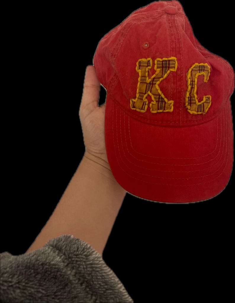 Pu&ograve; includere: Cappellino da baseball rosso con le lettere "KC" in tessuto a quadri giallo. Il cappellino &egrave; tenuto su uno sfondo nero. Ha una visiera curva e un cinturino in tessuto sul retro.