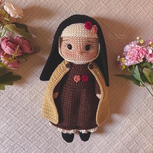 May include: A handmade crochet doll dressed as a nun, featuring a dark brown dress, a tan cape with two small buttons, and a black habit. The doll has a beige face with embroidered eyes, rosy cheeks, and small red and pink floral accents.