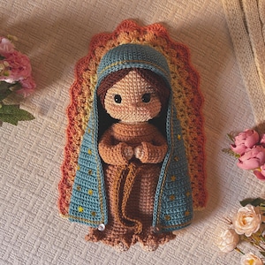 Puede incluir: Una muñeca de ganchillo que representa una figura con cabello castaño, vestida con un traje color canela y una capa azul con capucha adornada con pequeñas estrellas doradas. Detrás hay un halo de ganchillo semicircular en naranja, amarillo y rojo.