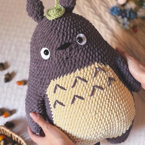 Puede incluir: Un muñeco de Totoro de peluche hecho a mano en tonos grises y crema. El muñeco presenta grandes ojos con bordes blancos, una pequeña hoja verde en la cabeza y una barriga color crema con marcas de chevrón gris oscuro. El muñeco es sostenido por manos.