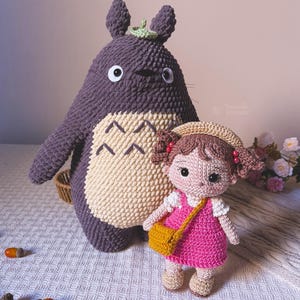 May include: Crochet dolls: a large purple Totoro and a girl in a pink dress. The Totoro has a cream belly and a green leaf. The girl doll has a yellow bag and a beige hat. Acorns are scattered in the foreground. The dolls are handmade.