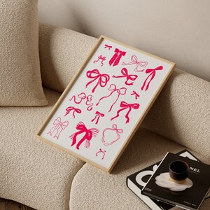 May include: A framed print featuring a collection of pink bow illustrations on a white background. The bows vary in style and size, with a simple, hand-drawn aesthetic. The frame is a light wood color, and the print is displayed on a beige sofa.