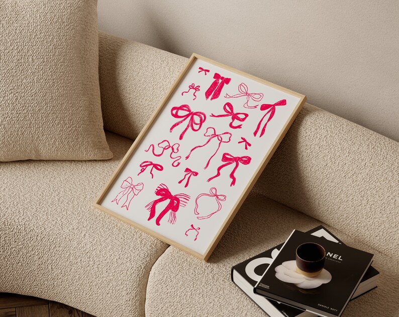 May include: A framed print featuring a collection of pink bow illustrations on a white background. The bows vary in style and size, with a simple, hand-drawn aesthetic. The frame is a light wood color, and the print is displayed on a beige sofa.