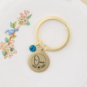 May include: Gold-tone keychain with a round charm that says "Oma" in cursive with a floral design. A small blue crystal dangles from the charm.