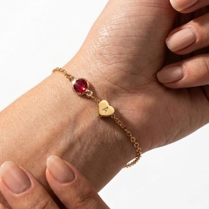 May include: Gold-toned bracelet featuring a round red gemstone and a heart-shaped charm with the letter "A". The delicate chain bracelet is worn on a wrist, showcasing its elegant design. The bracelet is a piece of jewelry.