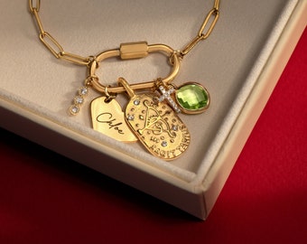 May include: A gold-toned chain necklace featuring a large oval carabiner clasp. Attached are four charms: a vertical bar with three clear stones, a heart-shaped tag engraved with the name Chloe, a Sagittarius zodiac coin, and a green stone cross.