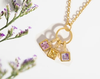 May include: A gold chain necklace with a circular pendant and three dangling square charms. The charms are set with pink, yellow, and purple gemstones.