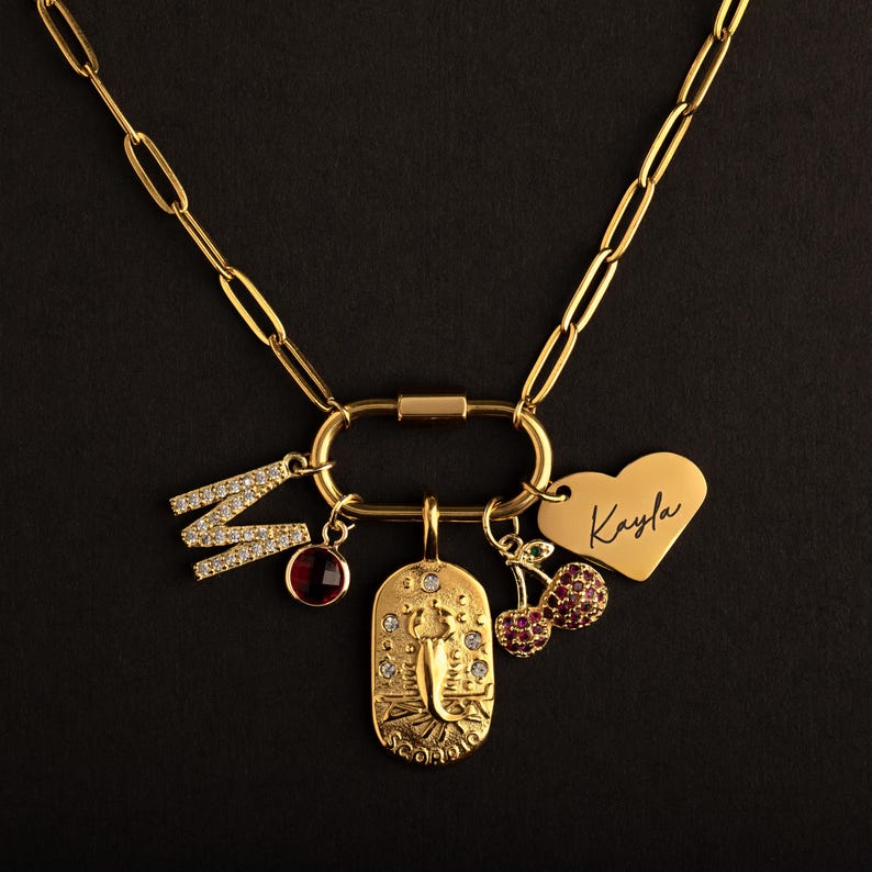May include: A gold-toned paperclip chain necklace featuring an oval carabiner clasp. Attached charms include a crystal-encrusted letter M, a round red stone, a rectangular Scorpio zodiac pendant, a cherry charm, and a heart tag engraved with Kayla.