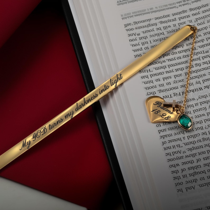May include: A gold bookmark with the inscription "My GOD turns my darkness into light" and a heart-shaped charm with the words "Psalms 18:28" and a green gemstone.