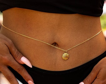 May include: A gold-colored body chain with a seashell pendant. The delicate chain is draped across the midriff, with the seashell charm centered. The skin tone is medium, and the background is blurred.