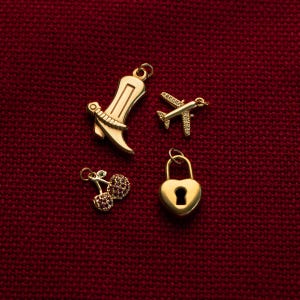 May include: Four gold-toned metal charms including a boot, a small airplane with clear stones, a pair of cherries with red stones, and a heart-shaped padlock with a keyhole. Each charm features a small circular loop for attachment to jewelry.