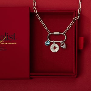 Silver paperclip carabiner necklace with customizable charms, featuring a blue birthstone, compass charm, and heart pendant, displayed in a red gift box. A meaningful and elegant gift option.