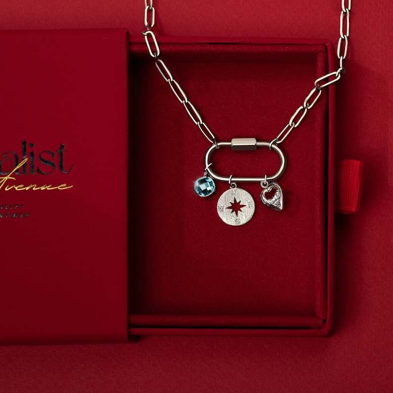 Silver paperclip carabiner necklace with customizable charms, featuring a blue birthstone, compass charm, and heart pendant, displayed in a red gift box. A meaningful and elegant gift option.