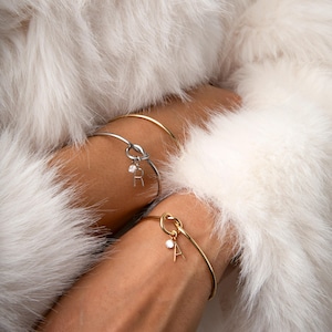Puede incluir: Dos pulseras delicadas, una plateada y otra dorada, cada una con un diseño de nudo y un pequeño dije con una letra y un acento brillante. Las pulseras se exhiben sobre un fondo de suave y blanca piel sintética.