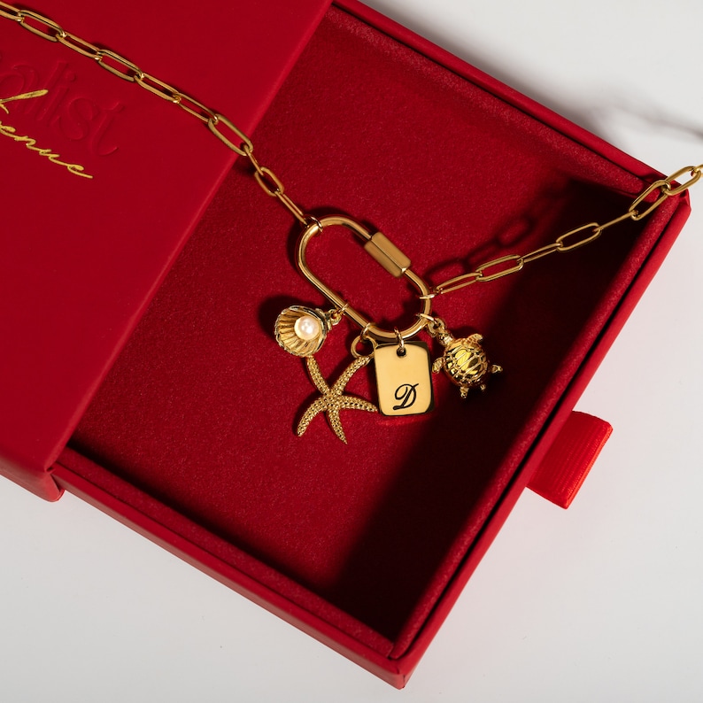 Gold paperclip carabiner necklace with customizable charms, including a pearl shell, starfish, initial tag with 'D,' and turtle charm, displayed in a red gift box. A unique, personalized accessory.