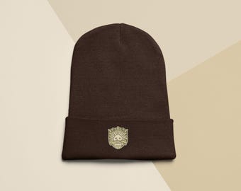 Puede incluir: Un gorro marrón oscuro con un emblema dorado. El emblema es un diseño de sol dentro de un escudo. El gorro está hecho de un material suave y tejido.