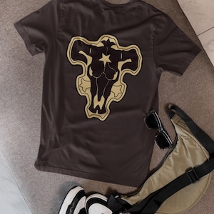 Puede incluir: Camiseta gris oscuro con un diseño de cabeza de toro en negro y dorado en la espalda. El diseño incluye una estrella. La camiseta se presenta con zapatillas deportivas en blanco y negro, gafas de sol y un bolso bandolera beige.