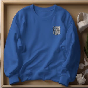May include: Royal blue sweatshirt with a crew neck and long sleeves. The sweatshirt features a white and gray emblem on the left chest. The emblem is a stylized design of wings and a crest. The sweatshirt is made of a soft material.