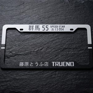 May include: A rectangular license plate frame featuring a two-tone design with a silver top section and a black bottom section. The top displays Japanese characters, the number 55, and the text SPEED STAR 13-954. The bottom features Japanese text and TRUENO.
