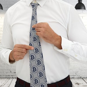 Könnte beinhalten: Eine graue Krawatte mit einem sich wiederholenden Muster aus blauen Bitcoin-Symbolen. Die Krawatte wird von einer Person in einem weißen Hemd und einer schwarzen Hose angepasst.