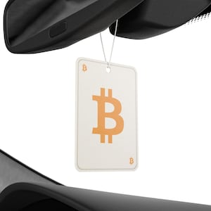 Puede incluir: Un ambientador de coche blanco con un gran símbolo de Bitcoin naranja. El ambientador está colgado del espejo retrovisor de un coche.