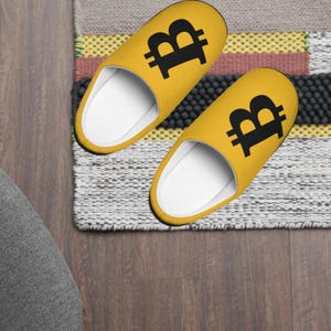 May include: A pair of yellow slippers with a black Bitcoin symbol on each. The slippers are on a gray and white rug.