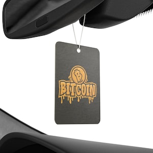 Puede incluir: Un ambientador rectangular negro para coche con la palabra "Bitcoin" en letras doradas estilizadas. El logotipo de Bitcoin está encima de la palabra. El ambientador cuelga de un cordón blanco, delante de un espejo retrovisor de coche.