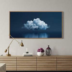 May include: A rectangular framed wall art piece featuring a single white cloud centred against a deep navy blue background, with a reflection of the cloud visible at the bottom. The artwork is displayed above a long, light-toned wooden sideboard.