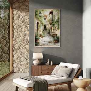 Op de afbeelding: Een ingelijste aquarel schildering toont een schilderachtige Europese straatsc&egrave;ne. Het kunstwerk toont een geplaveide steeg met gebouwen versierd met klimplanten en potbloemen. Een vintage straatlantaarn verlicht het pad en cre&euml;ert een serene sfeer.