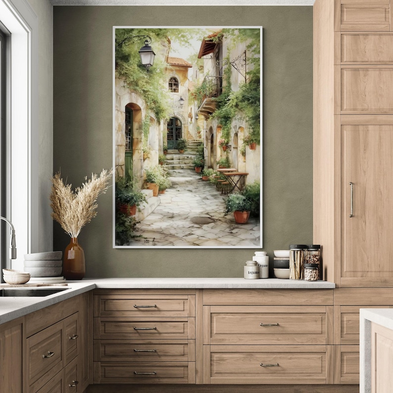 Op de afbeelding: Een ingelijste aquarelprint toont een Europese straatsc&egrave;ne. Het kunstwerk toont een geplaveide weg die zich slingert tussen gebouwen met groen en potplanten. Een vintage straatlantaarn verlicht de sc&egrave;ne en cre&euml;ert een charmante, ouderwetse sfeer.