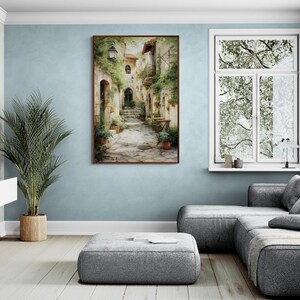 Op de afbeelding: Een ingelijste aquarel schildering toont een Europese dorpsstraat. Het kunstwerk toont een stenen pad, gebouwen met boogvormige deuren en weelderig groen. De schildering wordt tentoongesteld op een lichtblauwe muur, aangevuld met een grote potplant en een grijze hoekbank.