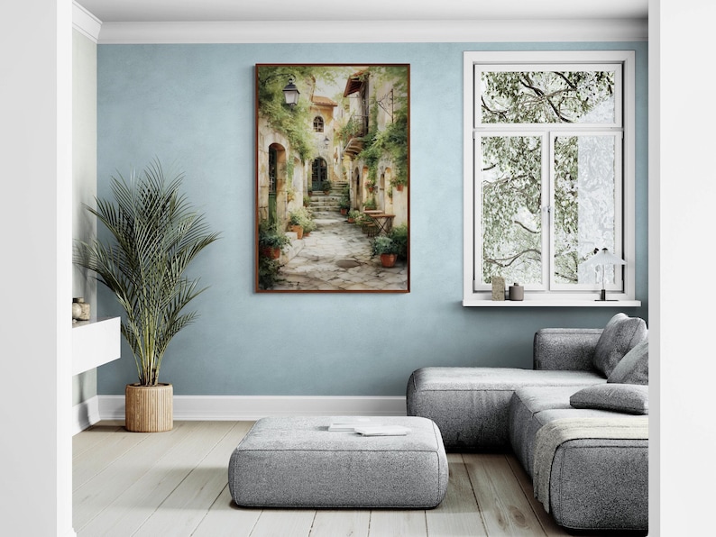 Op de afbeelding: Een ingelijste aquarel schildering toont een Europese dorpsstraat. Het kunstwerk toont een stenen pad, gebouwen met boogvormige deuren en weelderig groen. De schildering wordt tentoongesteld op een lichtblauwe muur, aangevuld met een grote potplant en een grijze hoekbank.